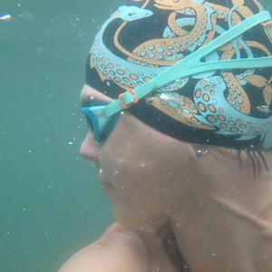 May include: A person wearing a black swim cap with an octopus design in teal and orange, and teal swim goggles. The person is underwater, with bubbles visible in the water.