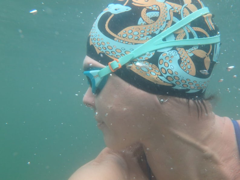 May include: A person wearing a black swim cap with an octopus design in teal and orange, and teal swim goggles. The person is underwater, with bubbles visible in the water.