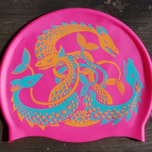 May include: A hot pink swim cap featuring a repeating pattern of stylized sea serpent illustrations in orange and teal. The sea serpents have detailed scales and fins, creating a vibrant and eye-catching design. The cap is made of a flexible material.