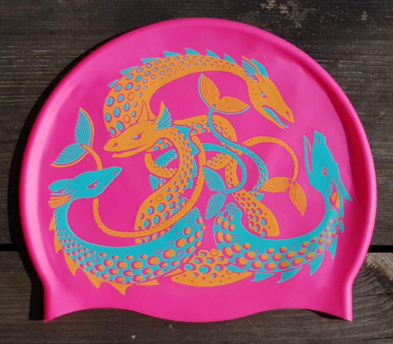 May include: A hot pink swim cap featuring a repeating pattern of stylized sea serpent illustrations in orange and teal. The sea serpents have detailed scales and fins, creating a vibrant and eye-catching design. The cap is made of a flexible material.