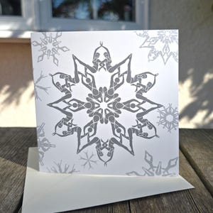 Puede incluir: Una tarjeta de felicitación blanca con un diseño de copo de nieve gris compuesto por figuras estilizadas en posturas de yoga. La tarjeta es cuadrada y viene con un sobre blanco. Se imprimen patrones de copos de nieve adicionales en la tarjeta.