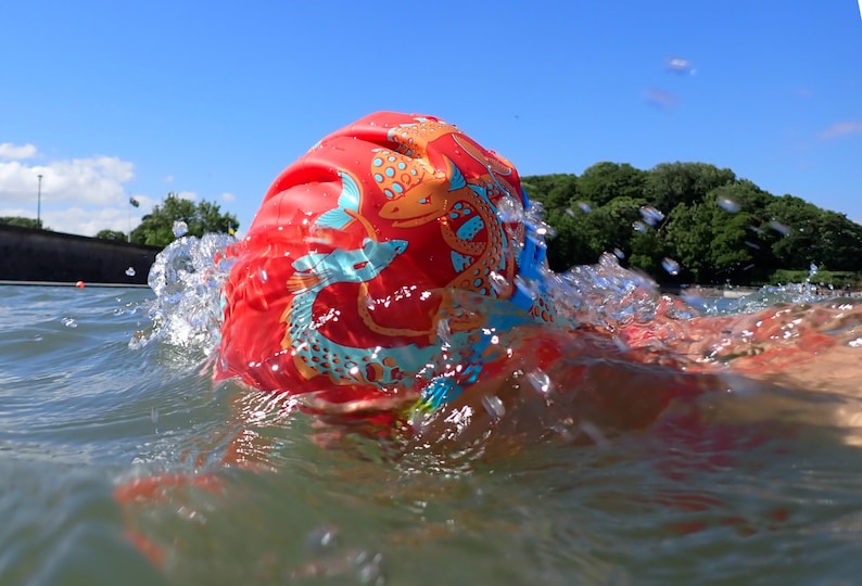 May include: A red swim cap with a koi fish design is partially submerged in water, creating a splash. The cap features orange and blue accents. The background shows a clear blue sky and green trees.