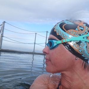 May include: A person wearing a black swim cap with an octopus design in orange and light blue, and teal swim goggles. The person is in the water, with a chain link fence and cloudy sky in the background.