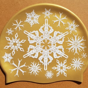May include: A gold-coloured swimming cap with a white snowflake design. The snowflakes are made up of stylised figures of people in a circle.