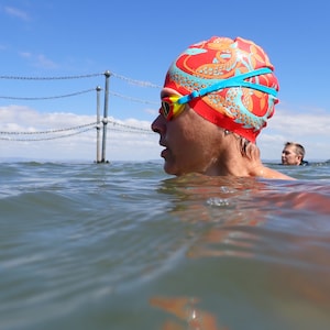 May include: A swimmer wearing a red swim cap with a dragon design and yellow and blue swim goggles. The swimmer is in the water, with a blue sky and a chain-link fence in the background. Another swimmer is visible in the distance.
