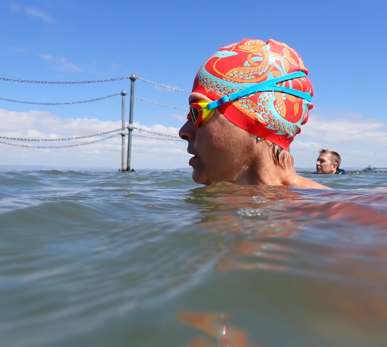 May include: A swimmer wearing a red swim cap with a dragon design and yellow and blue swim goggles. The swimmer is in the water, with a blue sky and a chain-link fence in the background. Another swimmer is visible in the distance.