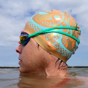 May include: A gold-colored swim cap with an orange and teal octopus design. The swimmer is also wearing green goggles and a green strap. The person is in the water, with a cloudy sky in the background.