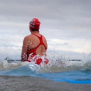 May include: A woman wearing a red swimsuit and a red and white swimming cap is swimming in the sea. She is surrounded by splashing water.