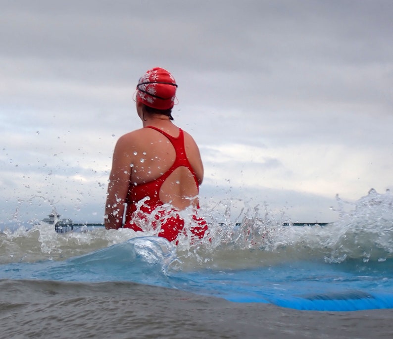 May include: A woman wearing a red swimsuit and a red and white swimming cap is swimming in the sea. She is surrounded by splashing water.