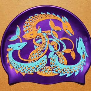 May include: Purple swim cap featuring a stylized design of intertwined sea serpents in orange and teal. The serpents have detailed scales and fins, creating a dynamic and eye-catching pattern. The cap is made of a smooth, shiny material.