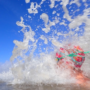 May include: A person wearing a colorful swim cap and goggles is partially submerged in water, with a large wave crashing over them. The bright blue sky contrasts with the white water, creating a dynamic and energetic scene.