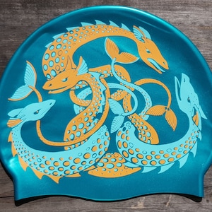 May include: Teal swim cap featuring a stylized design of orange and light blue sea serpents. The serpents are intertwined, with detailed scales and fins. The cap is made of a shiny, smooth material.