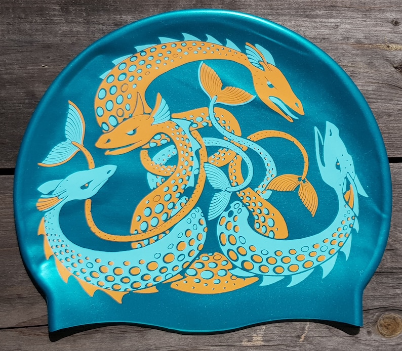 May include: Teal swim cap featuring a stylized design of orange and light blue sea serpents. The serpents are intertwined, with detailed scales and fins. The cap is made of a shiny, smooth material.