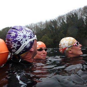 May include: Three people wearing swimming caps and goggles are in a body of water. The caps are decorated with snowflake designs. The person in the foreground is wearing a purple cap with white snowflakes, the person in the middle is wearing an orange cap with white snowflakes, and the person in the background is wearing a gold cap with white snowflakes.