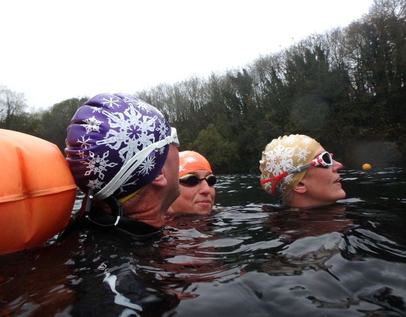 May include: Three people wearing swimming caps and goggles are in a body of water. The caps are decorated with snowflake designs. The person in the foreground is wearing a purple cap with white snowflakes, the person in the middle is wearing an orange cap with white snowflakes, and the person in the background is wearing a gold cap with white snowflakes.