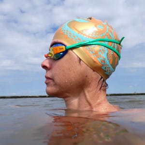 May include: A person wearing swimming goggles and a gold and teal swim cap with an octopus design. The goggles have a green strap and the brand name "TYR" is visible. The person is in the water, with a cloudy sky in the background.