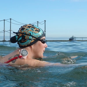 Op de afbeelding: Een zwemmer met een zwarte badmuts met een oranje en blauwgroen ontwerp, een zwarte zwembril en een roze sporthorloge. De zwemmer bevindt zich in het water, met een hek en een pier op de achtergrond.