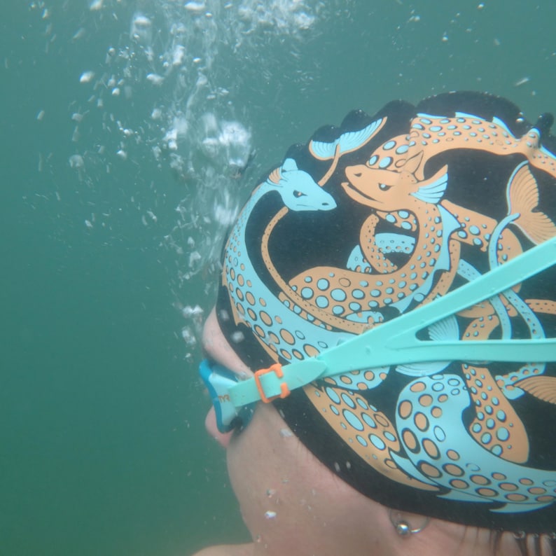 May include: A black swim cap with a sea serpent design in shades of orange and light blue. The cap is worn with light blue swim goggles. The image is taken underwater with bubbles in the background.