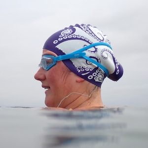 May include: A purple and white swim cap featuring a repeating octopus tentacle pattern, paired with light blue swimming goggles. The goggles have clear lenses and a blue strap that wraps around the head, worn over the patterned swim cap.