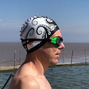 Könnte beinhalten: Eine weiß-schwarze Badekappe mit einem detaillierten Oktopus-Tentakel-Muster und einer kleinen orangefarbenen Fischgrafik. Die Kappe wird mit einer leuchtend grünen und schwarzen Schwimmbrille getragen, die den Markennamen HUUB am Rahmen trägt.