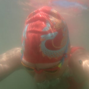 May include: A person wearing a red swim cap with a fish design and orange swim goggles underwater. The cap features a light blue and orange fish pattern. The image is taken from a first-person perspective, with the person's arms visible.