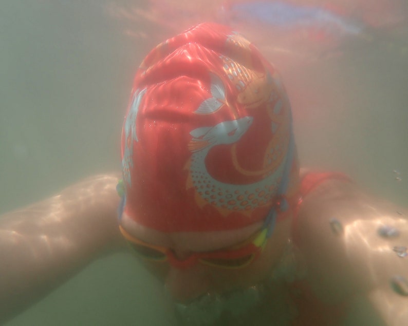 May include: A person wearing a red swim cap with a fish design and orange swim goggles underwater. The cap features a light blue and orange fish pattern. The image is taken from a first-person perspective, with the person's arms visible.