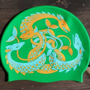May include: A bright green swim cap featuring a repeating design of stylized sea serpents in orange and light blue. The serpents are intertwined, creating a dynamic and eye-catching pattern. The cap is made of a flexible material.