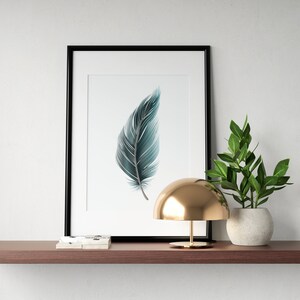 May include: A framed print of a teal blue feather on a white background. The print is on a shelf with a gold lamp and a potted plant.