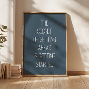 Könnte beinhalten: Ein gerahmter Druck mit dem Text "THE SECRET OF GETING AHEAD IS GETTING STARTED" in Weiß auf einem dunkelblauen Hintergrund. Der Rahmen hat eine hellbraune Farbe. Der Druck lehnt an einer Wand, mit Büchern und einer Pflanze sichtbar.