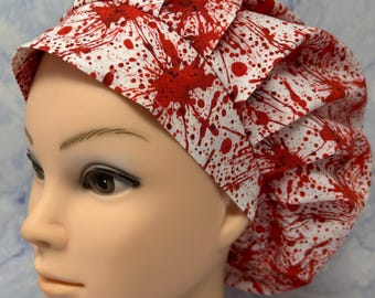 May include: A white surgical scrub cap with a repeating pattern of red blood splatters. The cap is made of fabric and has a pleated design. The cap is designed to cover the head and hair.