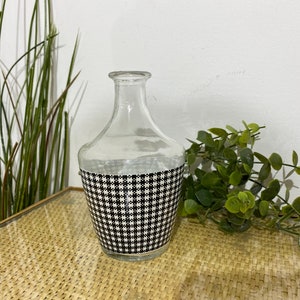 May include: A clear glass vase with a black and white checked pattern around the middle. The vase is sitting on a woven surface.
