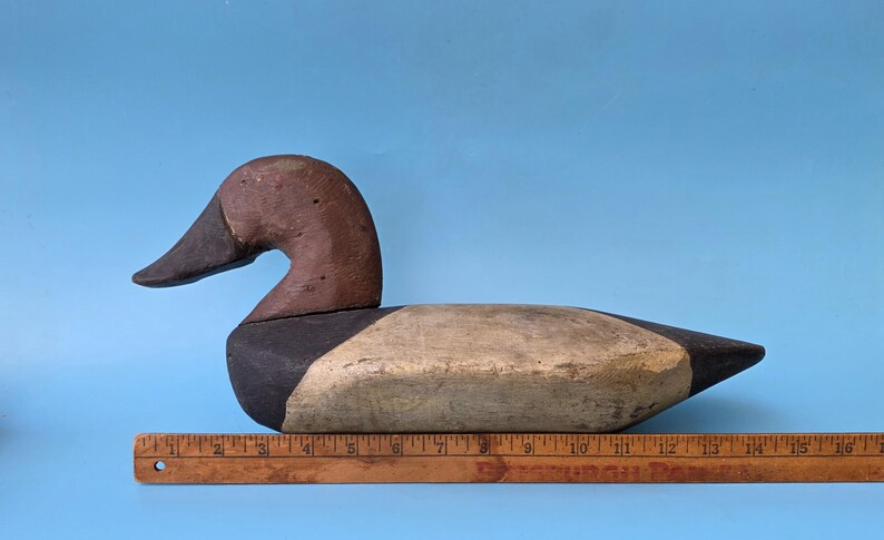 K&ouml;nnte beinhalten: Ein h&ouml;lzerner Entenk&ouml;der im Vintage-Stil mit braunem Kopf und schwarzem Schnabel. Der K&ouml;rper ist in Schwarz-, Beige- und Wei&szlig;t&ouml;nen bemalt. Der K&ouml;der ist etwa 30 cm lang und liegt auf einem Holzlineal vor blauem Hintergrund.