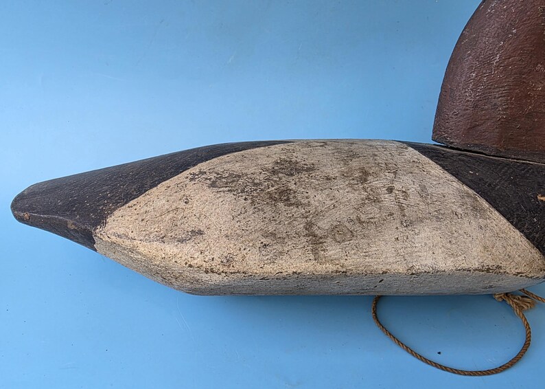 K&ouml;nnte beinhalten: Ein Vintage-Entenk&ouml;der, bemalt in Braun-, Schwarz- und Cremet&ouml;nen. Der K&ouml;der hat ein verwittertes Aussehen mit sichtbarer Textur und Abnutzung. Ein Seil ist an der Unterseite befestigt. Der K&ouml;der ist vor einem blauen Hintergrund platziert.
