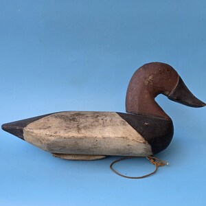 K&ouml;nnte beinhalten: Ein Vintage-Entenk&ouml;der, bemalt in Braun-, Schwarz- und Cremet&ouml;nen. Die Ente hat einen gebogenen Hals und einen detaillierten Schnabel. Ein Lederband ist befestigt. Der K&ouml;der ist vor einem einfarbigen blauen Hintergrund platziert.