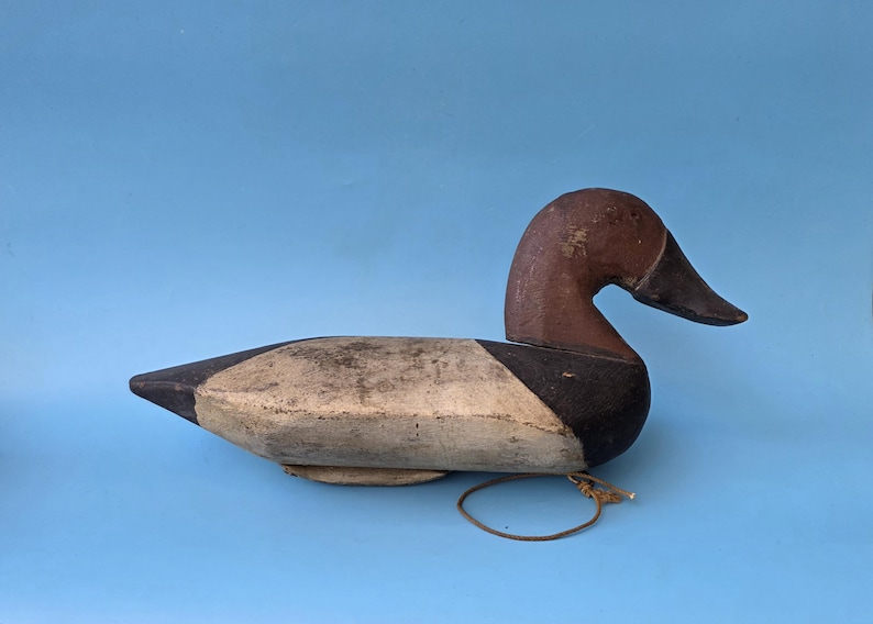K&ouml;nnte beinhalten: Ein Vintage-Entenk&ouml;der, bemalt in Braun-, Schwarz- und Cremet&ouml;nen. Die Ente hat einen gebogenen Hals und einen detaillierten Schnabel. Ein Lederband ist befestigt. Der K&ouml;der ist vor einem einfarbigen blauen Hintergrund platziert.