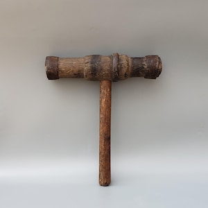 May include: A vintage wooden mallet with a cylindrical head and a long, slender handle. The head is dark brown with visible wear, and the handle is a lighter shade of brown. The tool is likely used for carpentry or woodworking.