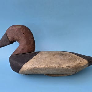 K&ouml;nnte beinhalten: Ein Vintage-Entenk&ouml;der aus Holz. Die Ente hat einen braunen Kopf und einen schwarzen Schnabel. Der K&ouml;rper ist in Beige und Schwarz bemalt und hat ein verwittertes Aussehen. Er befindet sich vor einem hellblauen Hintergrund.
