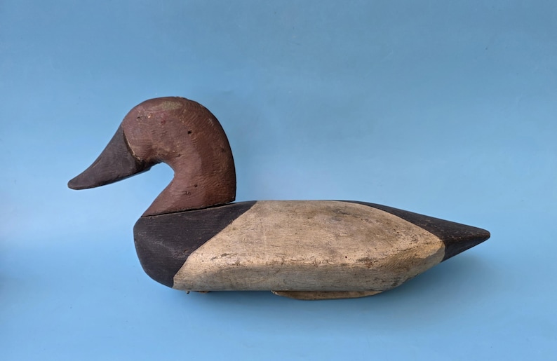 K&ouml;nnte beinhalten: Ein Vintage-Entenk&ouml;der aus Holz. Die Ente hat einen braunen Kopf und einen schwarzen Schnabel. Der K&ouml;rper ist in Beige und Schwarz bemalt und hat ein verwittertes Aussehen. Er befindet sich vor einem hellblauen Hintergrund.