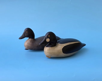 May include: Two wooden duck decoys with black heads, beaks, and backs, and tan and white bodies. The ducks are on a light blue background. The decoys are likely used for hunting or as decorative items.