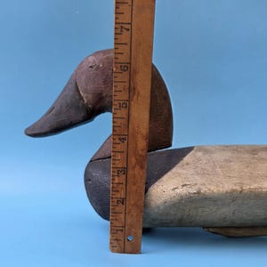 K&ouml;nnte beinhalten: Ein Vintage-Holzentenk&ouml;der mit braunem Kopf, schwarzem Schnabel und cremefarbenem K&ouml;rper. Der K&ouml;der ist etwa 18 cm lang, mit einem Holzlineal zur Ma&szlig;stabsbestimmung. Die Ente befindet sich vor einem blauen Hintergrund.