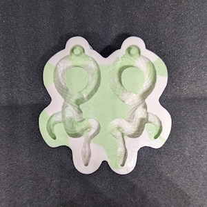 Puede incluir: Un molde de silicona blanco con dos cavidades en forma de serpiente. El molde está diseñado para hacer pendientes de resina.