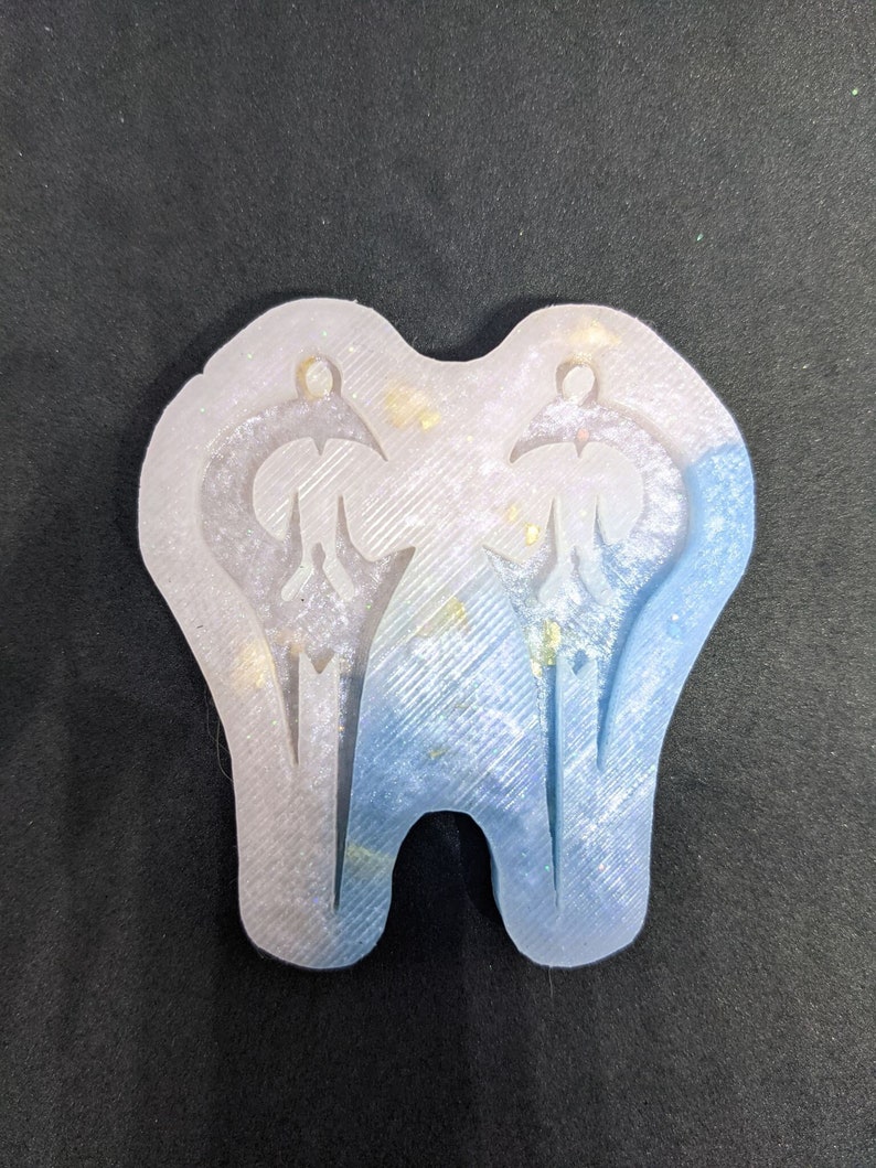 May include: A clear silicone mold with two tooth-shaped cavities. The mold is filled with blue and white glitter.
