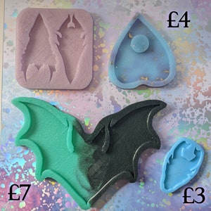 May include: Four silicone moulds for resin crafts. The moulds are shaped like a bat, a heart with a circle inside, a feather, and a teardrop. The moulds are green, black, blue, and purple.