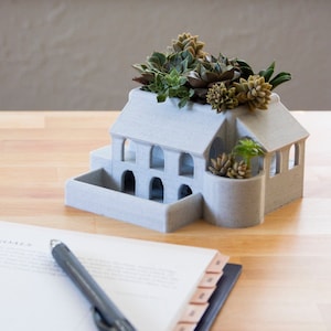 May include: A white ceramic house-shaped planter with a green succulent plant growing out of the top. The planter has a textured surface and several small windows.