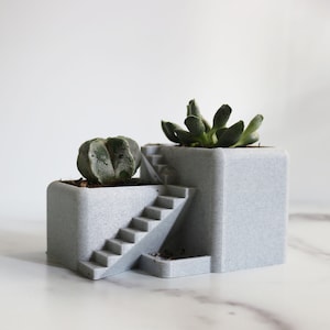 Puede incluir: Un macetero de hormigón gris con una escalera integrada, que presenta dos suculentas. El macetero tiene una superficie texturizada y está diseñado para contener varias plantas.