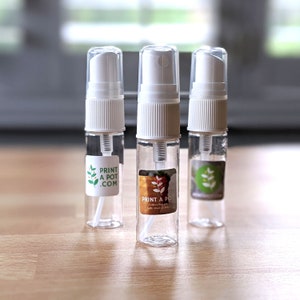 May include: Three clear spray bottles with white tops and labels that read "PRINT A POT .COM". The labels feature a plant graphic. The bottles are on a light brown surface.