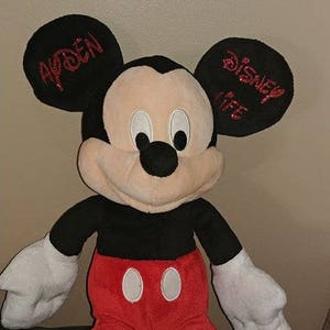 Może przedstawiać: Pluszowa zabawka Mickey Mouse w kolorze czarnym i czerwonym z dużymi ż&oacute;łtymi stopami. Uszy są czarne z czerwonym błyszczącym tekstem, kt&oacute;ry na jednym uchu m&oacute;wi "Disney Life", a na drugim "Ayden".