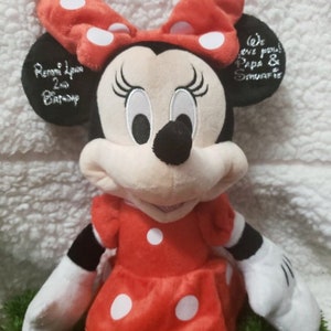 Może przedstawiać: Pluszowa zabawka Minnie Mouse w czerwonym kolorze w białe kropki z czarną kokardką. Zabawka ma wyszyte rzęsy i uśmiechnięte usta. Zabawka ma spersonalizowany napis na uchu, kt&oacute;ry brzmi "Remmi Lou 2nd Birthday".