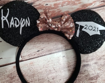 Pode incluir: Tiara de orelhas de rato com lantejoulas pretas com um laço de lantejoulas em ouro rosa. Uma orelha tem o nome "Kadyn" em escrita branca, e a outra orelha apresenta um gráfico de chapéu de formatura e o ano "2021" em branco.