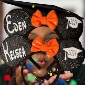 May include: Black and white graduation cap Mickey Mouse ears with orange bows and the names "Eden" and "Kelsea" written on them. The ears also have the year "2024" written on them.